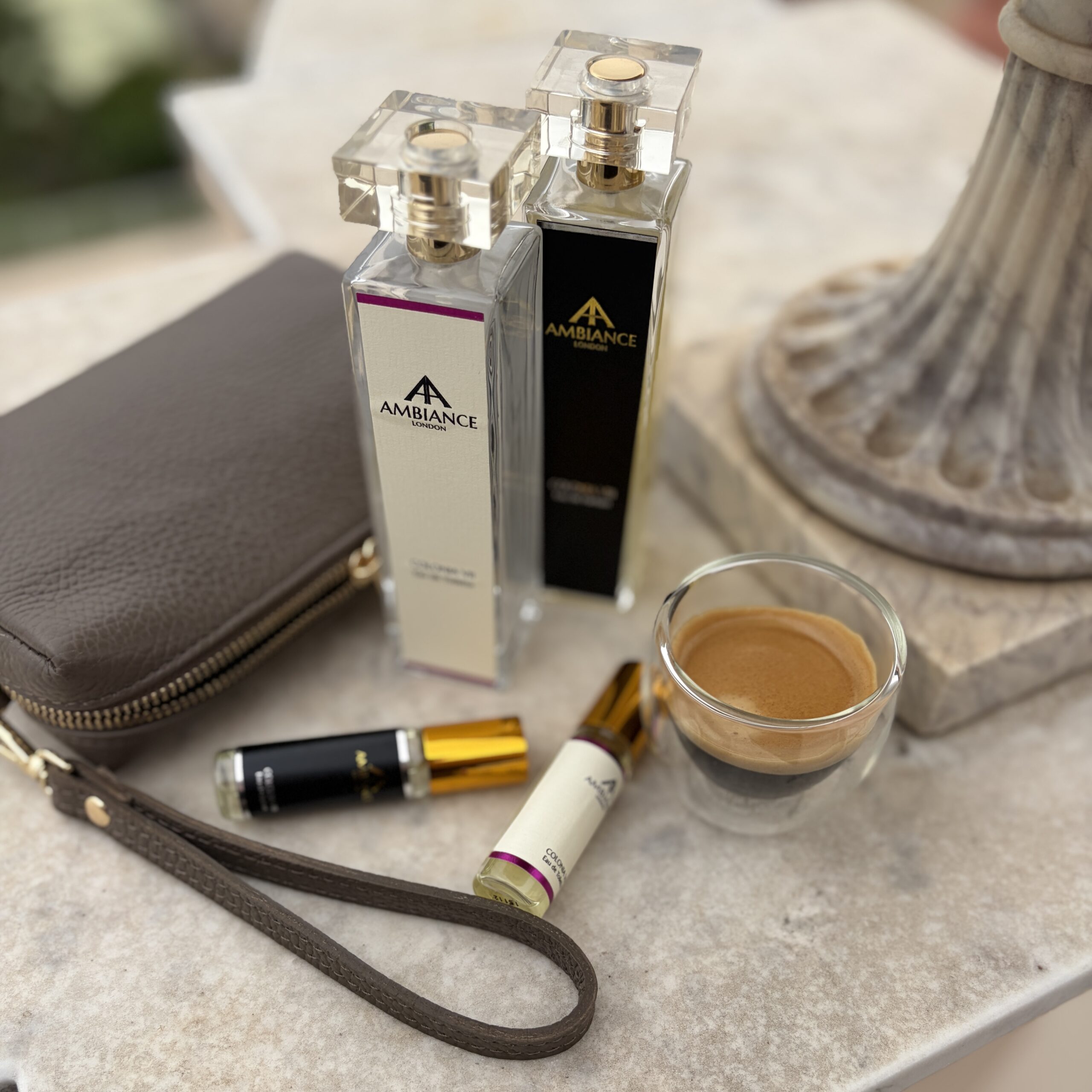 Taupe Bellezza leather pochette with Ambiance London perfumes, travel sprays and espresso on a marble tabletop.