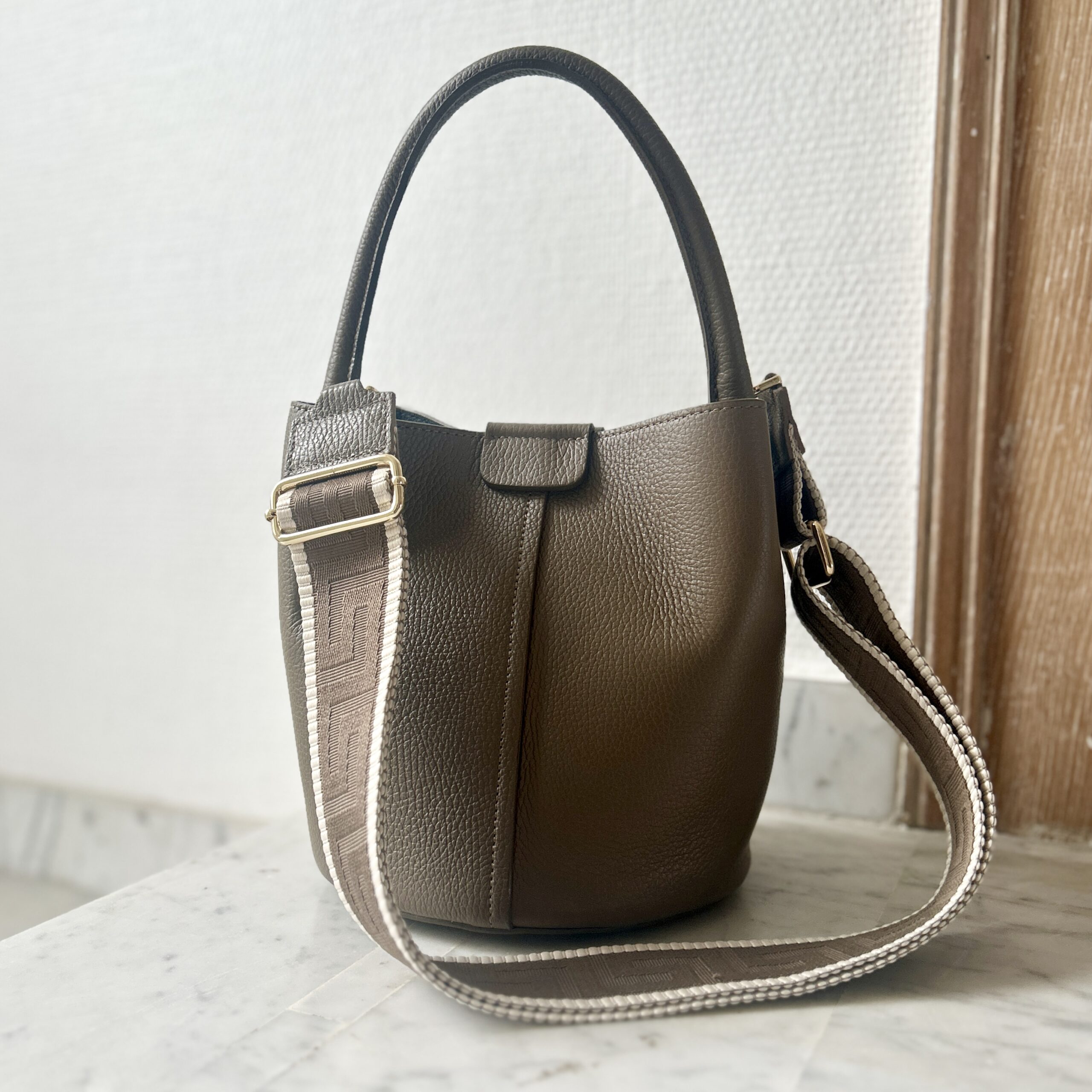 Taupe pebbled leather Dalu bucket bag with a matching top handle, detachable taupe and cream woven shoulder strap, and gold hardware on a marble surface.