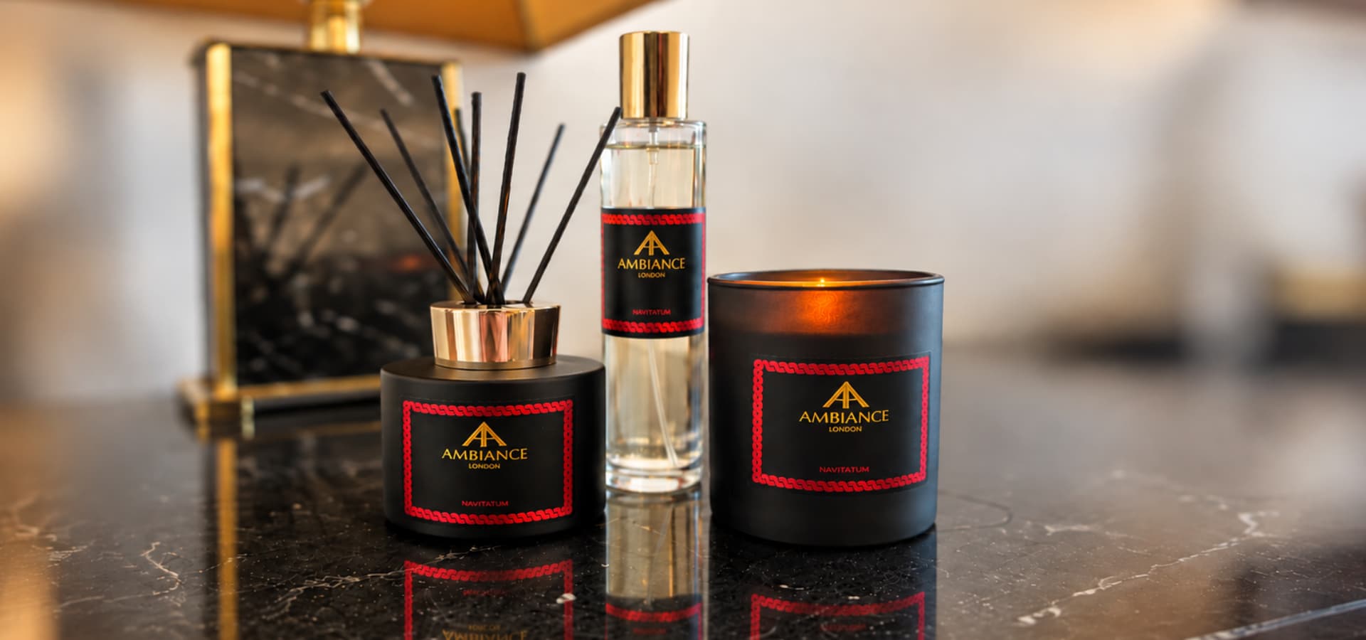 Navitatum luxury home fragrance collection by Ambiance London, featuring a lit black glass candle, a matte black reed diffuser with gold collar and black reeds, and a clear glass room spray with a gold cap, styled on black marble with warm brass accents.