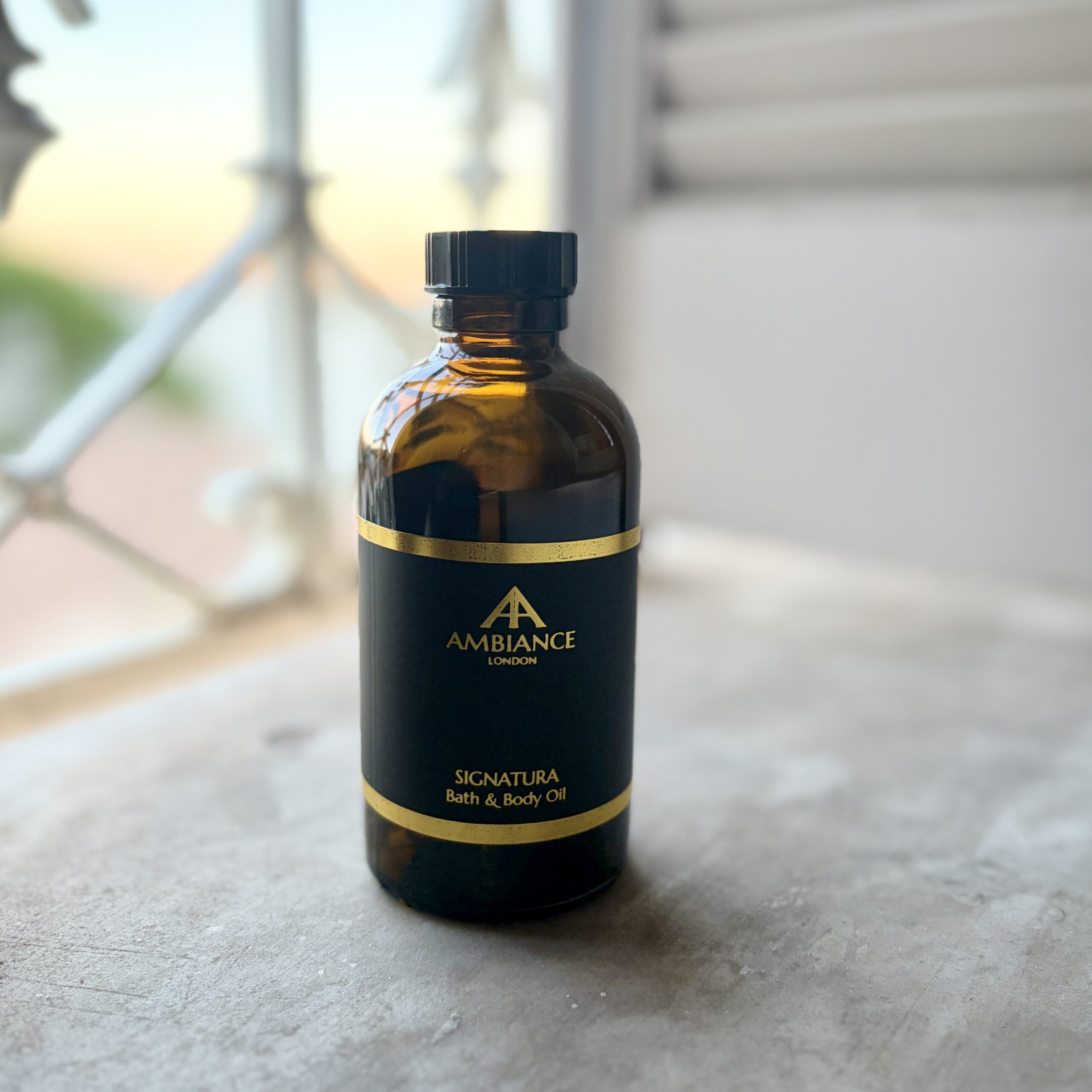 Signatura Bath & Body Oil 200ml in an amber glass bottle with a black and gold label beside a softly lit window.