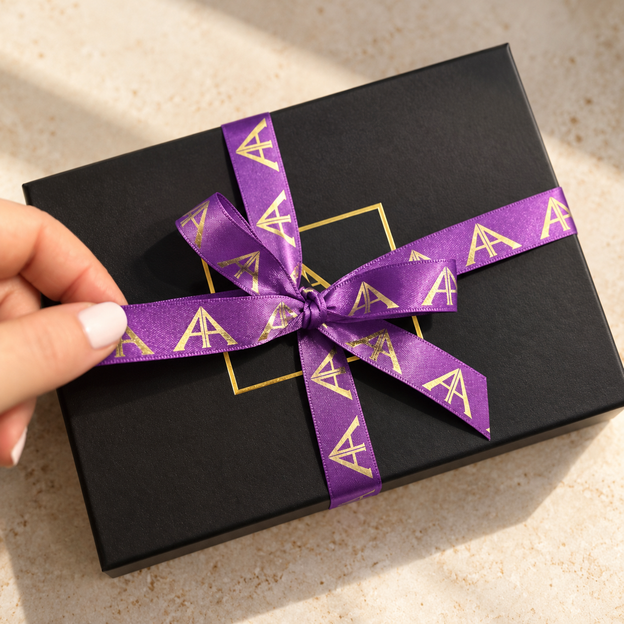 Black Ancienne Ambiance gift box wrapped with purple satin ribbon and gold logo, presented on a neutral stone background with a hand gently holding the bow.