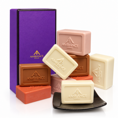 A collection of Ancienne Ambiance London soap bars in diverse colours, including white, pink, brown, and orange, arranged neatly beside a purple gift box. The scene conveys luxury and elegance.
