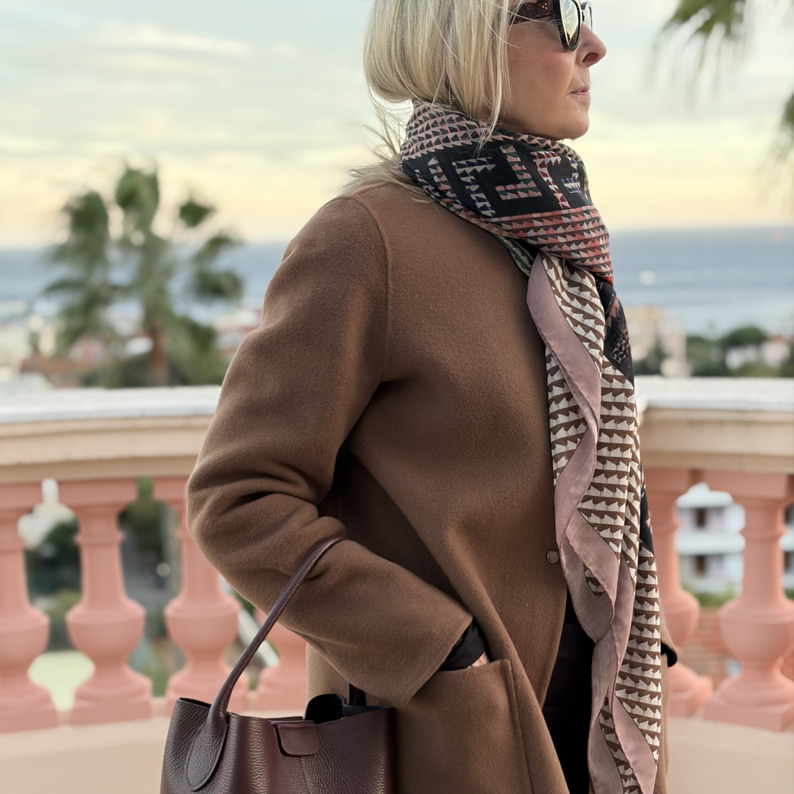 Model wearing the Greco Romana Travertine wool-silk scarf with a brown coat and carrying the Dalu Bucket Bag in Pompeian Plum.