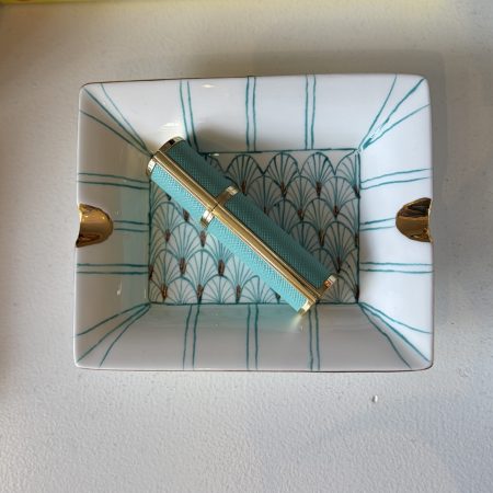 A turquoise perfume atomiser lays on a rectangle shaped porcelain tray with turquoise painted pattern and gold-plated accents.