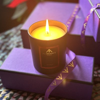 A lit Ancienne Ambiance London Navitatum Candle sits elegantly amongst a selection of purple gift boxes.
